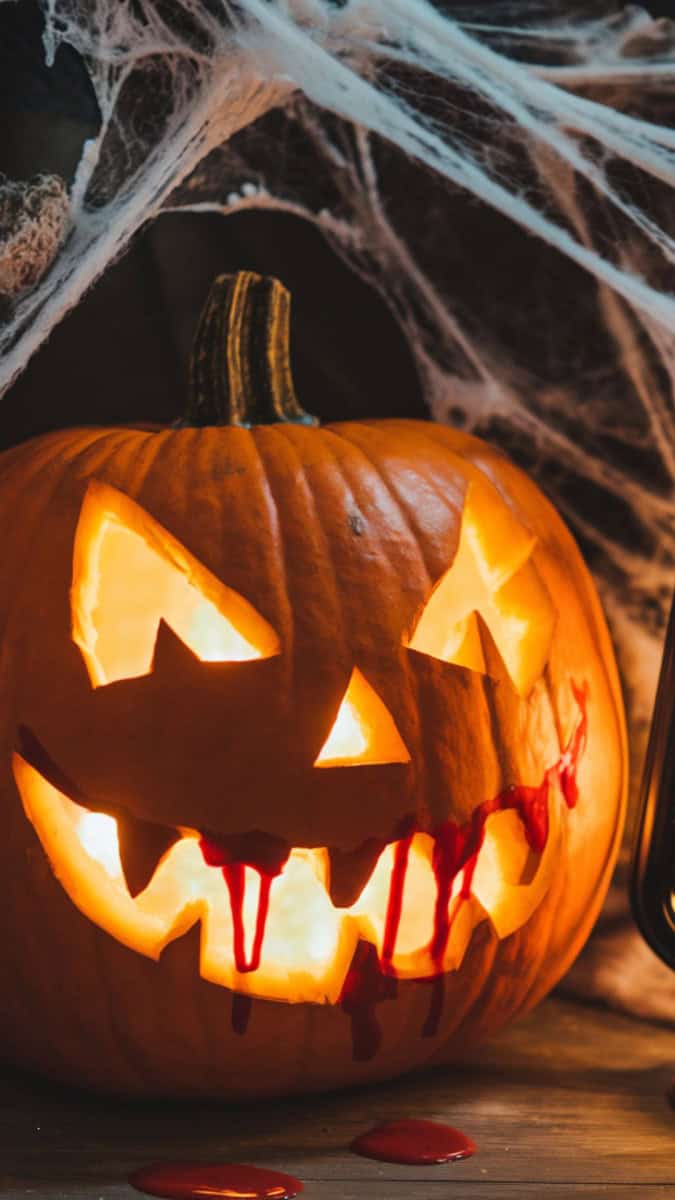 A carved pumpkin face with sharp plastic fangs and painted red “blood” dripping from the mouth. Styled with cobwebs, bats, and a glowing lantern beside it for spooky vibes.