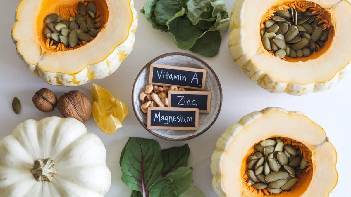A flat lay of halved white pumpkins, their seeds scooped into a small rustic bowl beside fresh leafy greens, nuts, and small chalkboard labels showing “Vitamin A,” “Zinc,” and “Magnesium.”