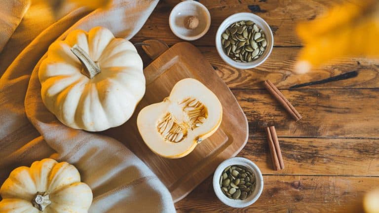 A cozy autumn kitchen scene with a rustic wooden table, a white pumpkin sliced open to reveal its pale interior, surrounded by small bowls of pumpkin seeds, cinnamon sticks, and nutmeg. Soft warm lighting with golden tones.