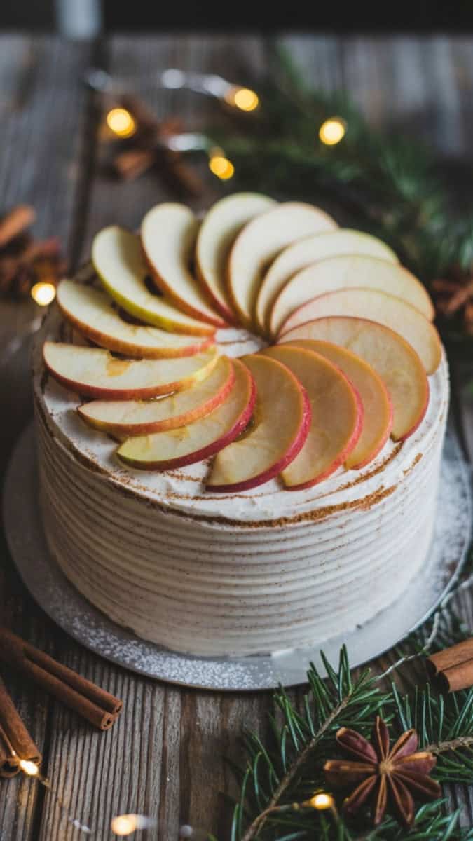 A rustic round cake frosted with cinnamon buttercream, decorated with thin apple slices arranged in a spiral on top, lightly dusted with powdered sugar. Styled on a wooden table with cinnamon sticks, pine sprigs, and golden fairy lights.