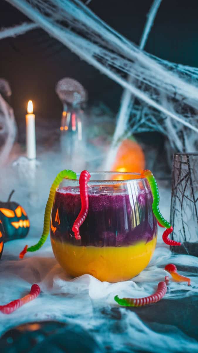 A spooky punch bowl filled with layered purple and yellow juice, gummy worms crawling over the rim. Styled with fog, spiderwebs, and Halloween glasses.