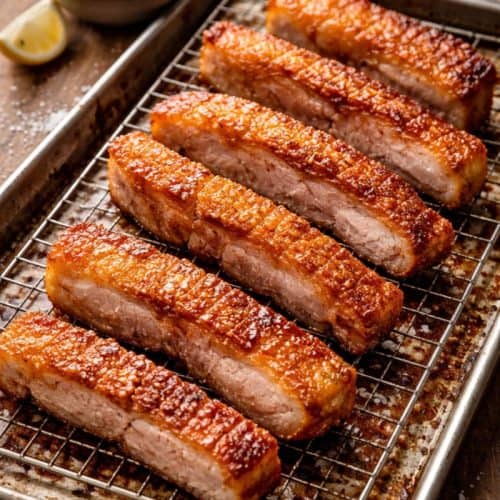 Crispy pork belly strips on a wire rack over a baking sheet, with golden crackling skin and rendered fat below, plus a bowl of flaky salt in the background.