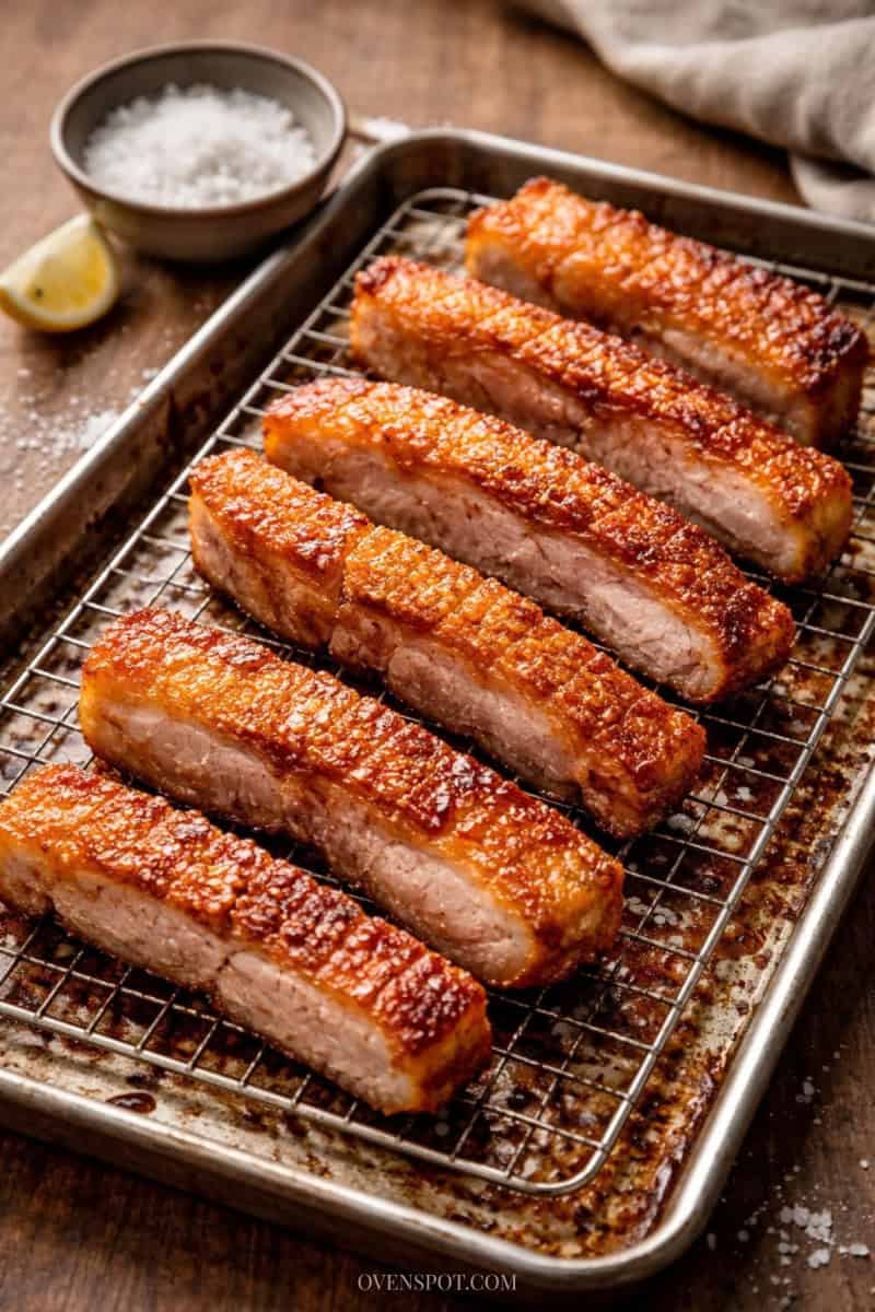 Crispy pork belly strips on a wire rack over a baking sheet, with golden crackling skin and rendered fat below, plus a bowl of flaky salt in the background.