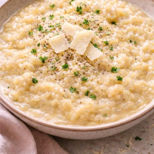 Creamy Parmesan risotto in a shallow white bowl, with a glossy, flowing texture and tender rice grains, topped with 3–4 shaved Parmesan pieces, cracked black pepper, and fresh parsle
