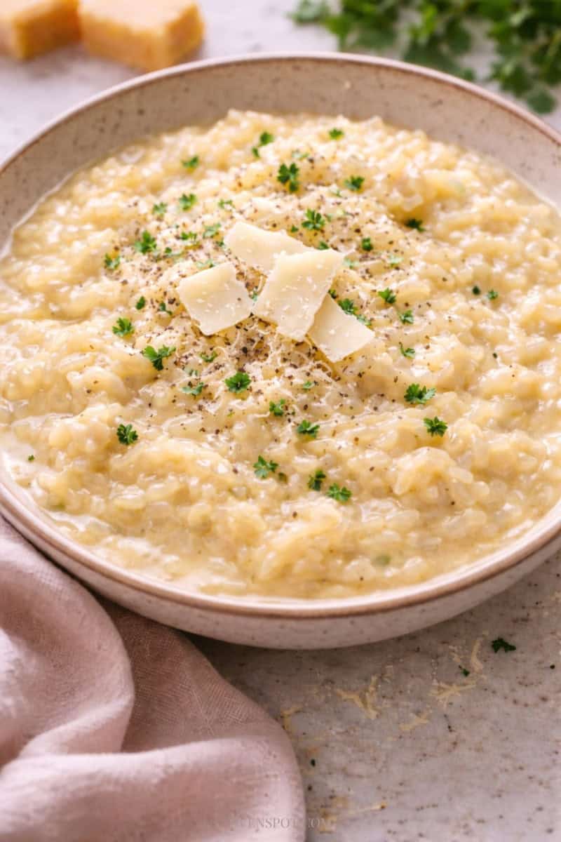 Creamy Parmesan risotto in a shallow white bowl, with a glossy, flowing texture and tender rice grains, topped with 3–4 shaved Parmesan pieces, cracked black pepper, and fresh parsle