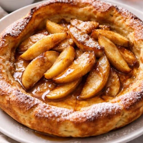 Apple cinnamon German pancake topped with sautéed apples, powdered sugar, and maple syrup on a white plate.