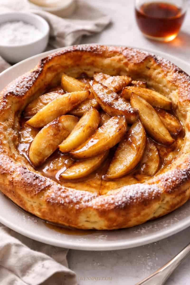 Apple cinnamon German pancake topped with sautéed apples, powdered sugar, and maple syrup on a white plate.