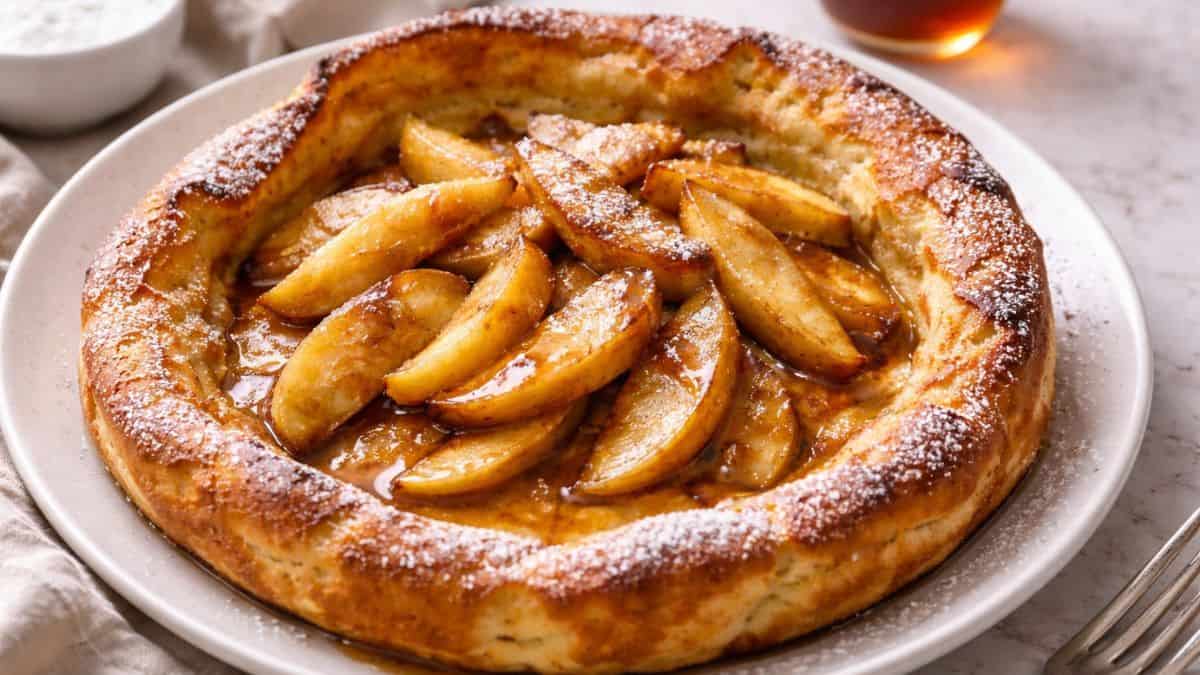 Apple cinnamon German pancake topped with sautéed apples, powdered sugar, and maple syrup on a white plate.