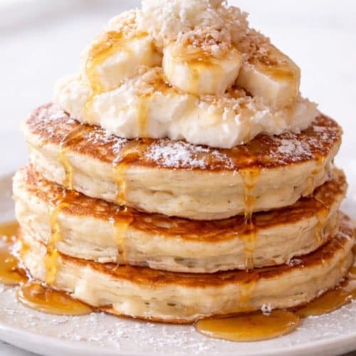 Sweet coconut cream pancakes stacked on a white plate with coconut cream drizzle, toasted coconut, and white chocolate curls.