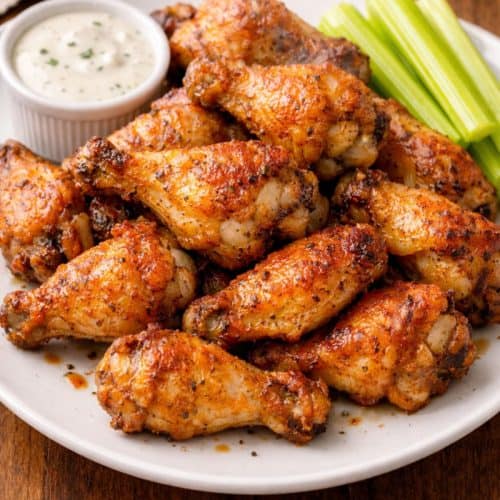 Crispy baked chicken wings piled on a white plate with ranch dipping sauce and celery sticks, golden-brown and crackly with visible seasoning.