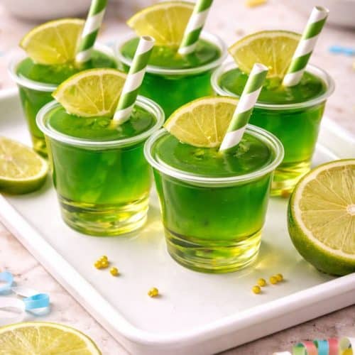 Six bright green lime jello shots in clear plastic cups topped with fresh lime wedges and striped paper straws, arranged on a white tray with sliced limes and colorful party decorations in the background.