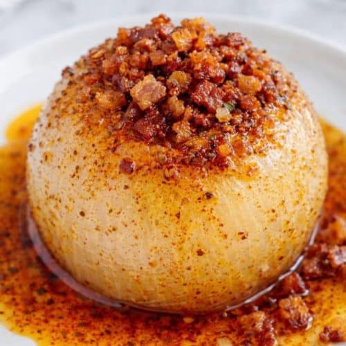 Whole onion boil on a small white plate with buttery spice sauce and crispy bacon