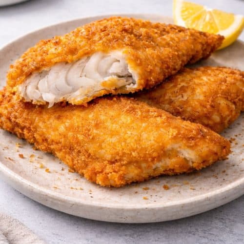 Crispy golden fried fish fillets on a speckled ceramic plate, with one fillet broken open to show flaky white fish, garnished with a lemon wedge on a modern light-gray background.