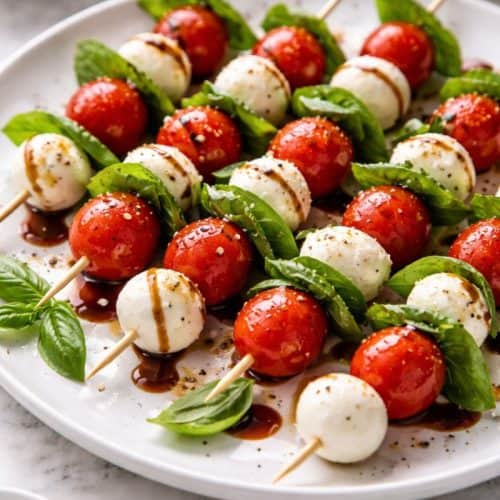 Caprese salad skewers on a white plate with cherry tomatoes, mozzarella, basil, and balsamic drizzle