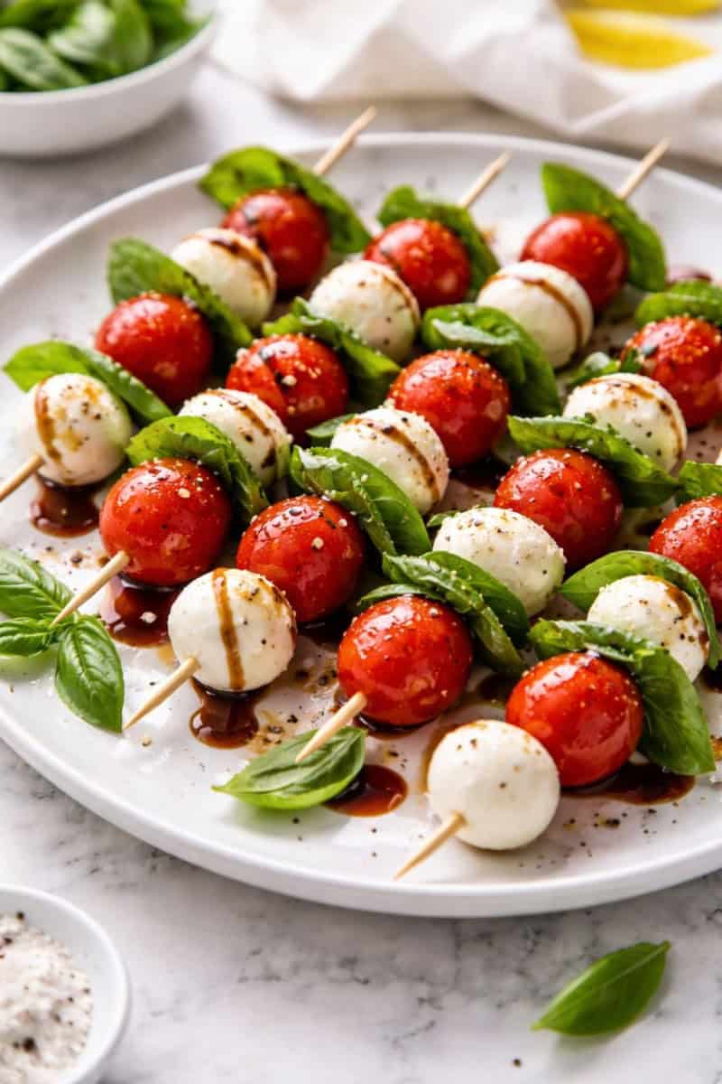 Caprese salad skewers on a white plate with cherry tomatoes, mozzarella, basil, and balsamic drizzle
