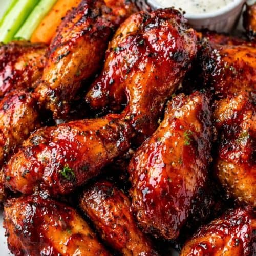 Crispy baked BBQ chicken wings piled on a white plate with glossy caramelized sauce, served with celery sticks, carrot sticks, and a ramekin of creamy dipping sauce.