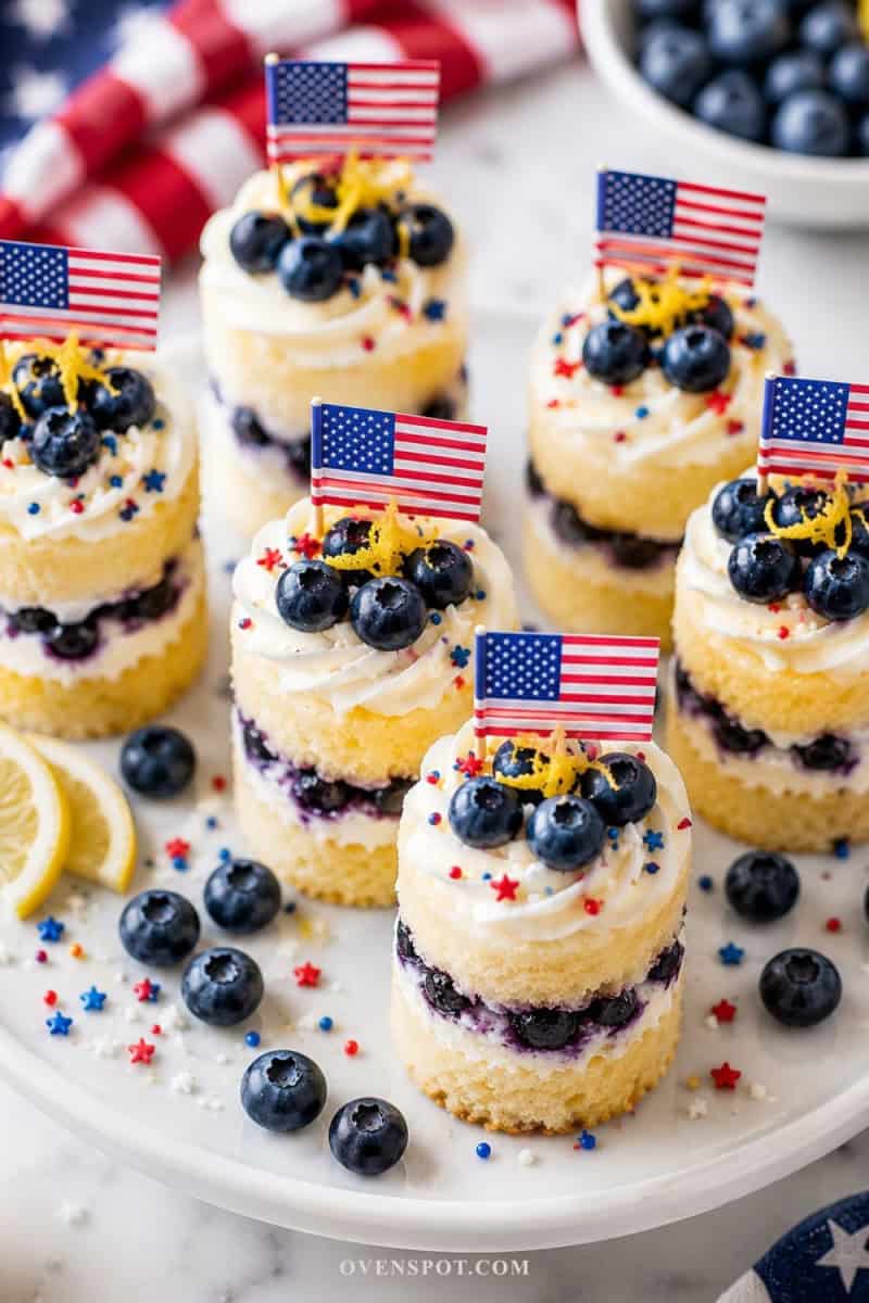 Lemon blueberry mini layer cakes topped with whipped frosting, fresh blueberries, lemon zest, patriotic sprinkles, and mini American flags