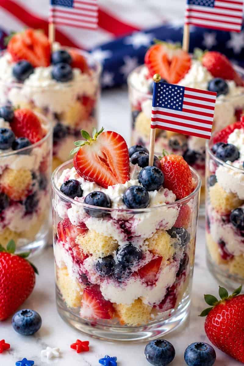 Patriotic mini berry shortcake trifles layered with whipped cream, strawberries, blueberries, and sponge cake in clear dessert glasses topped with mini American flags