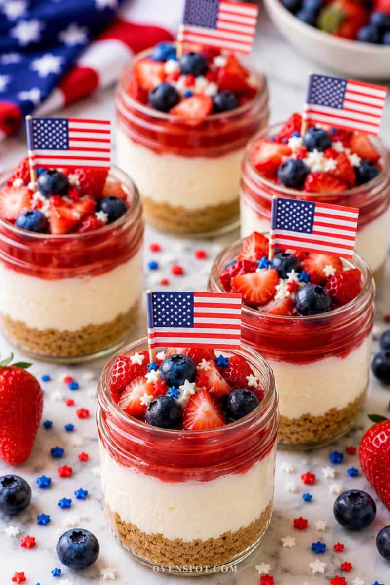 Patriotic strawberry cheesecake mini jars with graham cracker crust, creamy cheesecake filling, fresh strawberries, blueberries, and mini American flags