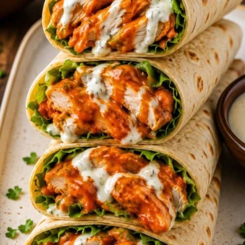 buffalo chicken wraps sliced on a white plate showing layers of spiced chicken, lettuce, avocado and cheese, with ranch dressing on the side