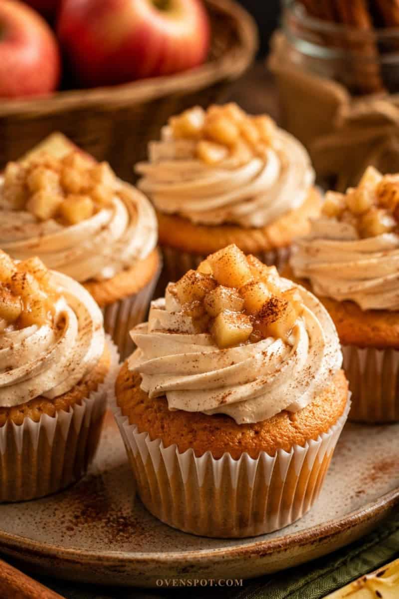 Apple pie cupcakes topped with cinnamon frosting and warm apple pie filling, styled in a cozy autumn kitchen setting with apples and cinnamon sticks in the background.