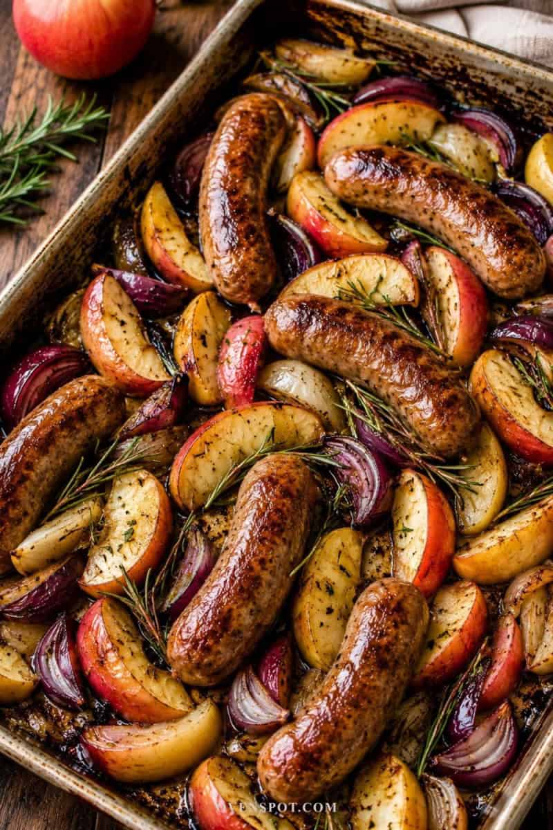 Sheet pan sausage and apples roasting together with red onions and fresh rosemary, featuring caramelized edges and golden browned sausage in warm rustic lighting.