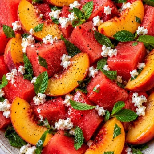 Fresh peach watermelon salad served in a large speckled ceramic bowl on a light marble surface. Juicy watermelon cubes and ripe peach wedges are tossed with crumbled feta, chopped mint, and cracked black pepper