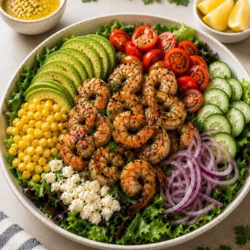 grilled shrimp salad served in a large white bowl with crisp mixed greens, sliced avocado, juicy cherry tomatoes, cucumber, sweet corn, thinly sliced red onion, and crumbled feta