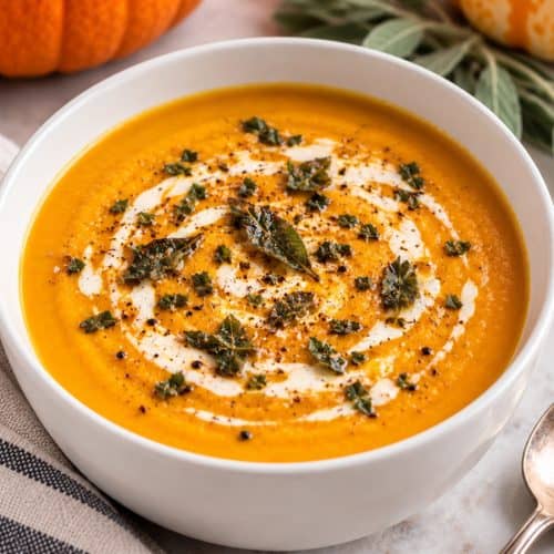 Creamy pumpkin soup in a white bowl, topped with crushed butter-crisped sage leaves, a swirl of cream, and cracked black pepper.